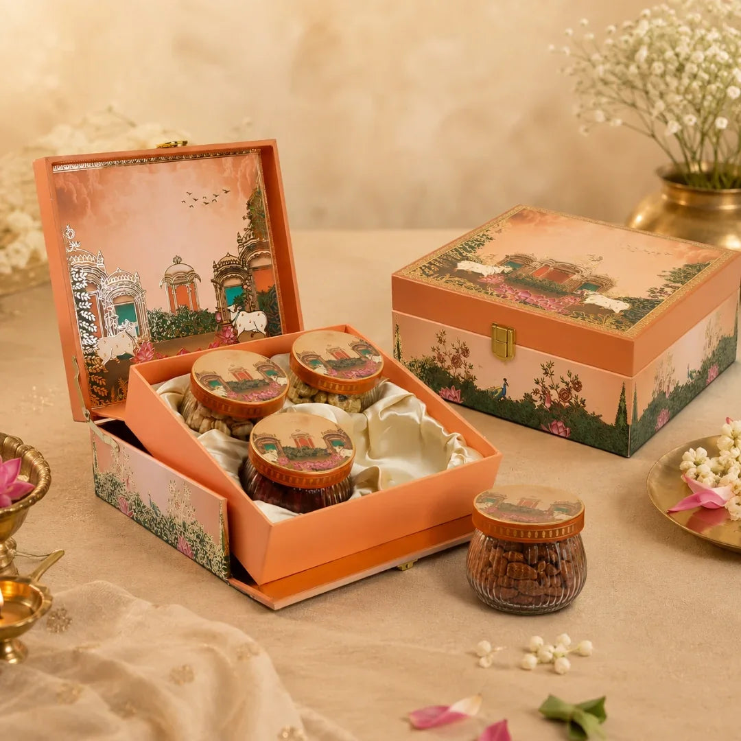 An open decorative gift box containing three small jars of snacks or sweets, with the matching closed box beside it on a beige fabric surface. The packaging has a warm peach-orange border with illustrated floral and architectural scenes, and the setup includes soft props like flowers and a brass vessel in a styled indoor setting.