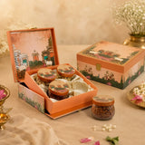 An open decorative gift box containing three small jars of snacks or sweets, with the matching closed box beside it on a beige fabric surface. The packaging has a warm peach-orange border with illustrated floral and architectural scenes, and the setup includes soft props like flowers and a brass vessel in a styled indoor setting.