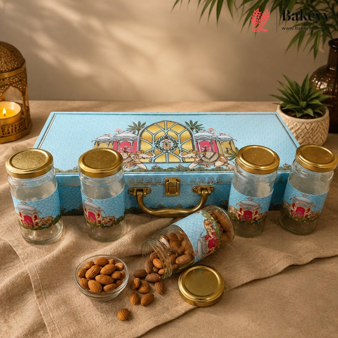 A set of five clear glass jars with gold lids and decorative blue labels, arranged in front of a matching blue illustrated storage case on a beige fabric surface. Almonds are scattered in front, with a candle, lantern, and potted plant in the background.