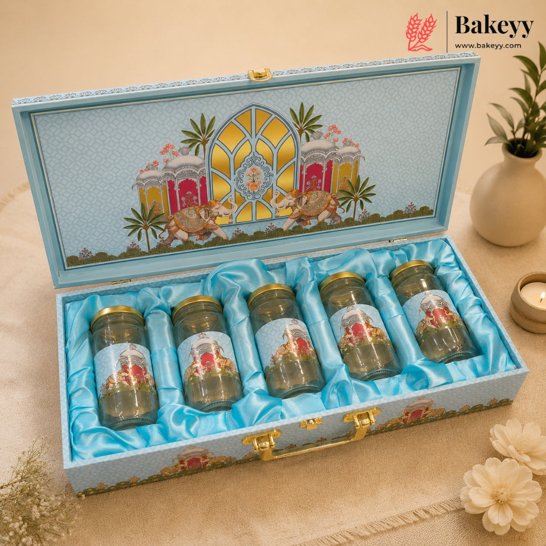 An open decorative gift box containing five small glass jars with gold lids, set on a beige surface with flowers and a plant in the background. The box lid is decorated with a colorful architectural illustration, and the interior is lined with light blue satin.