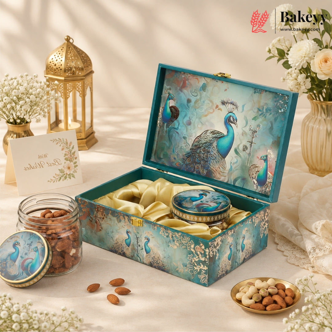 An ornate teal and gold gift box with peacock artwork on the lid, shown open with satin lining and a matching round tin inside. The box is styled on a cream fabric surface with flowers, a lantern, nuts, and a card.