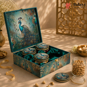 An ornate teal and gold gift box with peacock artwork, shown open to reveal three round jars of sweets or nuts lined up inside. Additional loose pieces and one open jar sit beside the box on a styled tabletop with warm lighting, decorative flowers, and a gold-toned screen in the background.
