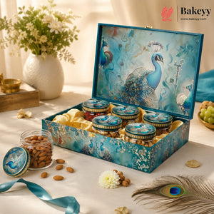 A decorative blue gift box with peacock artwork on the lid, filled with small round jars and displayed on a draped table with almonds, a flower, and a peacock feather. The Bakeyy logo appears in the top right corner.
