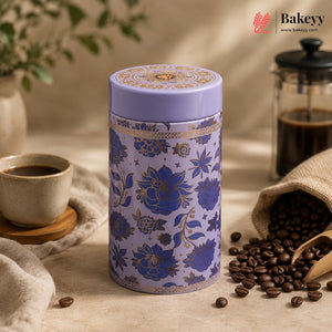 A cylindrical tin canister with a lavender purple lid and base, decorated with a blue and gold floral pattern, set on a beige fabric surface with coffee beans, a cup of coffee, and a burlap sack in a warm, styled setting. The Bakeyy brand logo is visible in the top right corner.