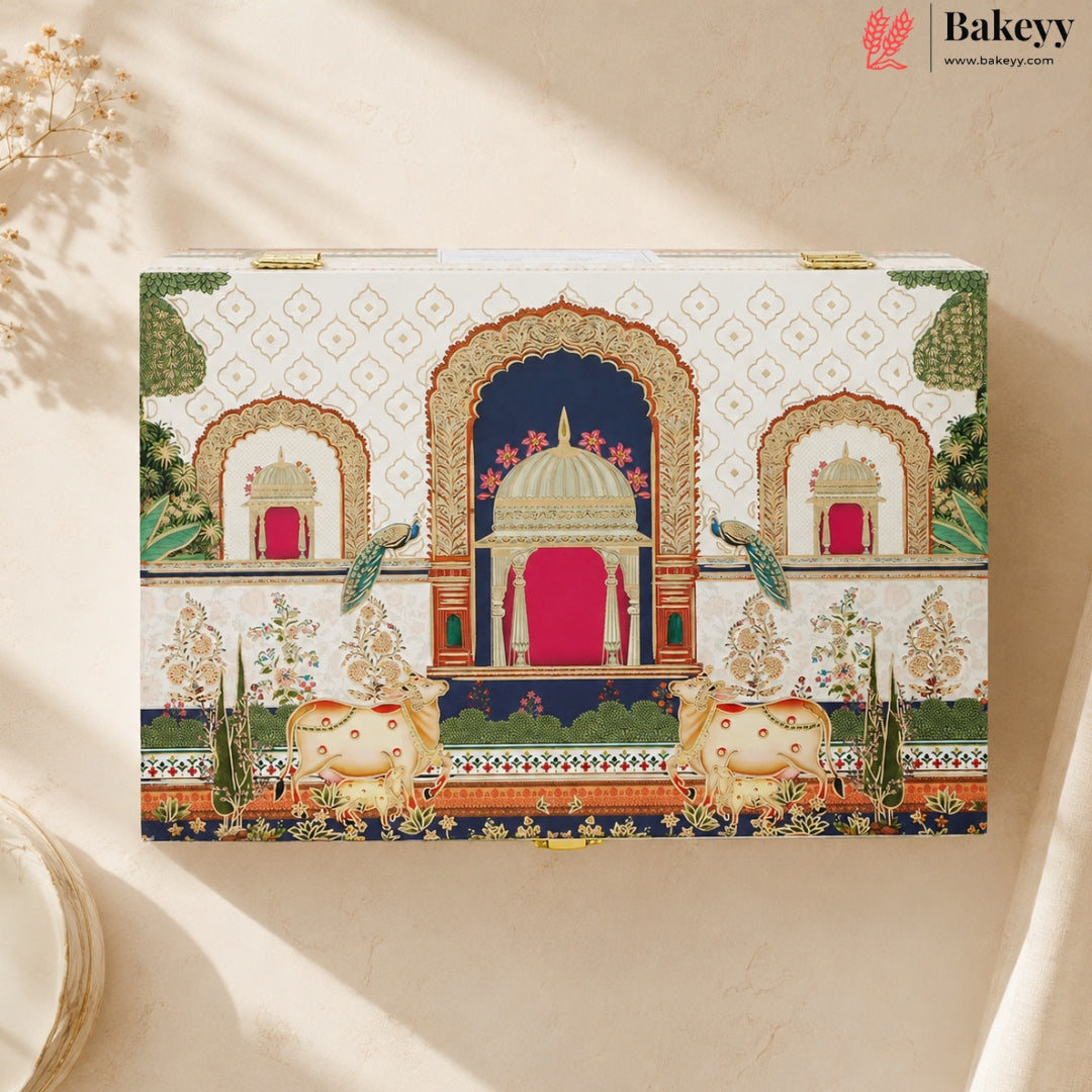 A rectangular decorative gift box with an ornate Indian architectural illustration, featuring arches, peacocks, elephants, and a red central door, photographed on a warm beige surface with soft shadows. The Bakeyy logo appears in the top right corner.
