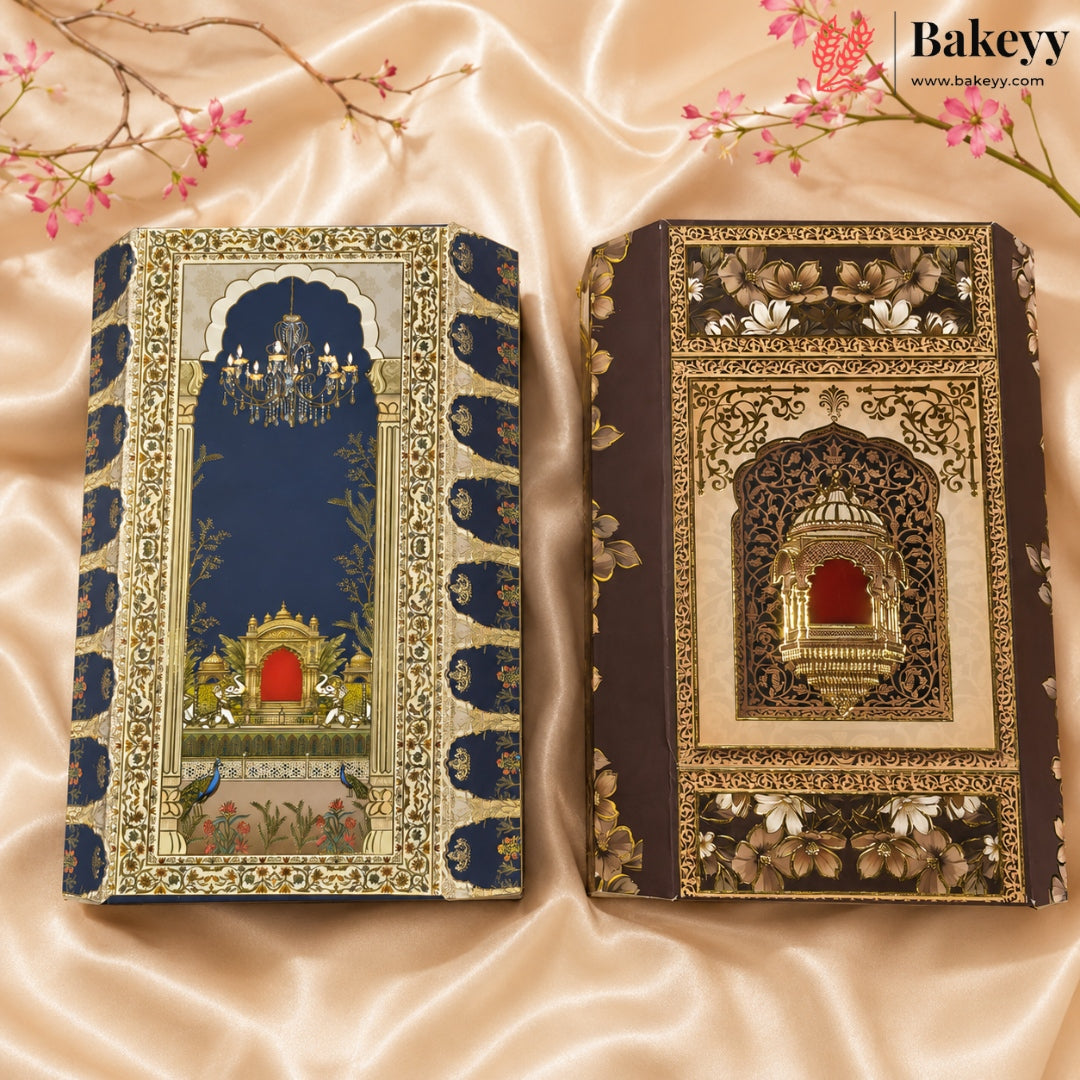 Two ornate decorative book-shaped cakes with intricate floral and architectural designs, displayed on a beige satin fabric background with cherry blossom branches. The left cake has a navy blue cover with gold and white detailing, and the right cake has a dark brown cover with gold and white detailing.