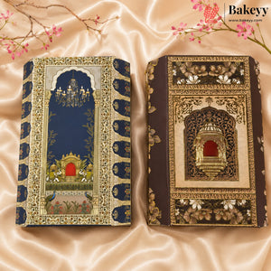 Two ornate decorative book-shaped cakes with intricate floral and architectural designs, displayed on a beige satin fabric background with cherry blossom branches. The left cake has a navy blue cover with gold and white detailing, and the right cake has a dark brown cover with gold and white detailing.