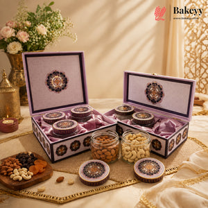 An elegant gift box set of baked goods from Bakeyy, featuring two ornate purple and gold boxes with glass jars of dates and nuts, styled on a table with a floral arrangement and candle.