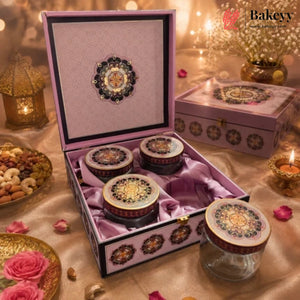 A pink decorative gift box set containing four glass jars with ornate gold and multicolor patterned lids, displayed on a draped satin surface with warm ambient lights and floral props. The Bakeyy brand logo appears in the top right corner.