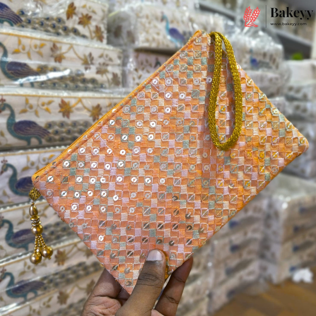 A hand holds a peach-colored clutch bag decorated with gold sequins and a gold wrist strap, with a matching gold tassel charm. The background shows stacks of packaged patterned fabric bags.
