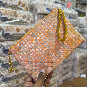 A hand holds a peach-colored clutch bag decorated with gold sequins and a gold wrist strap, with a matching gold tassel charm. The background shows stacks of packaged patterned fabric bags.