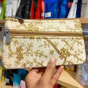 A hand holding a small zippered pouch with gold floral embroidery on a cream fabric background, set against a blurred store shelf backdrop.