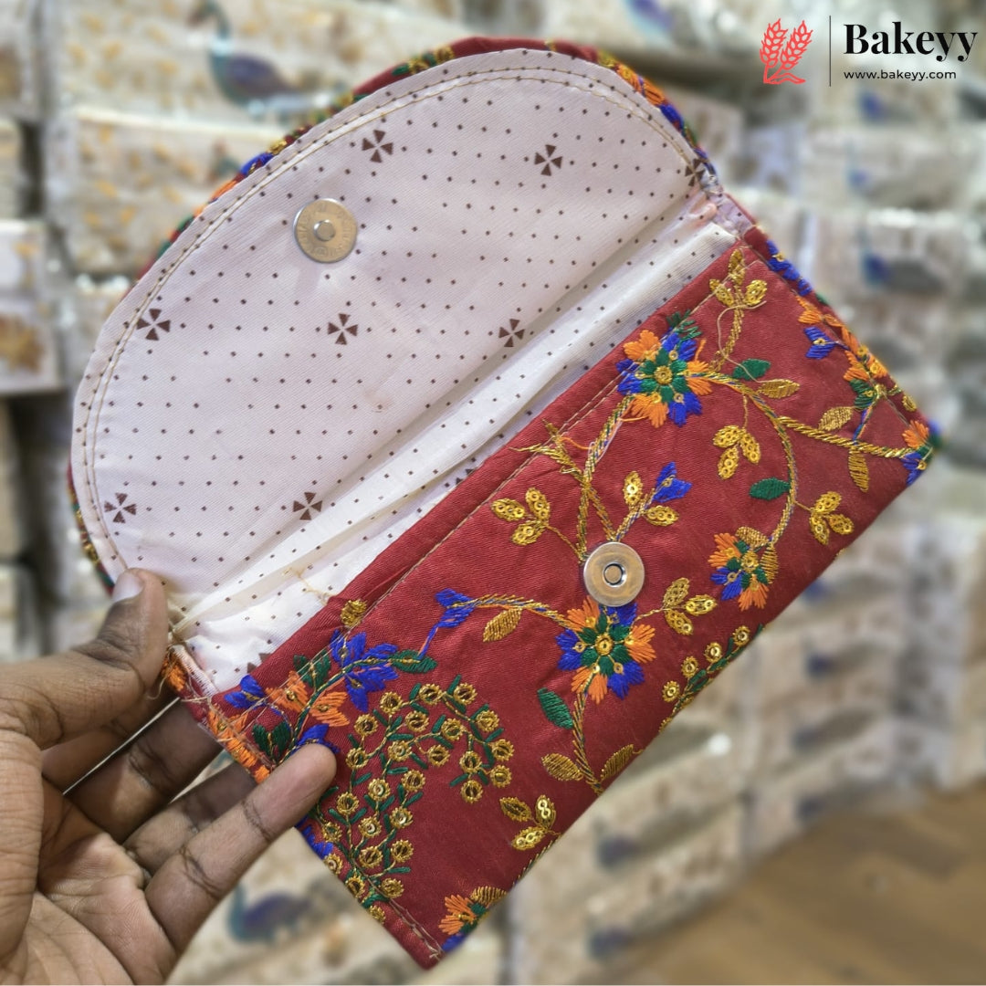 A handmade fabric clutch wallet held open by a hand, featuring a red floral embroidered exterior, a light pink dotted interior lining, and gold-tone snap buttons. The background shows a blurred tiled surface with a Bakeyy watermark logo in the top right.