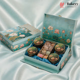 A decorative turquoise gift box with a lotus pond illustration, shown open to reveal four round pastries with floral lids and a central tray of assorted chocolates. The closed matching box sits beside it on draped satin fabric, with the Bakeyy logo in the top right corner.