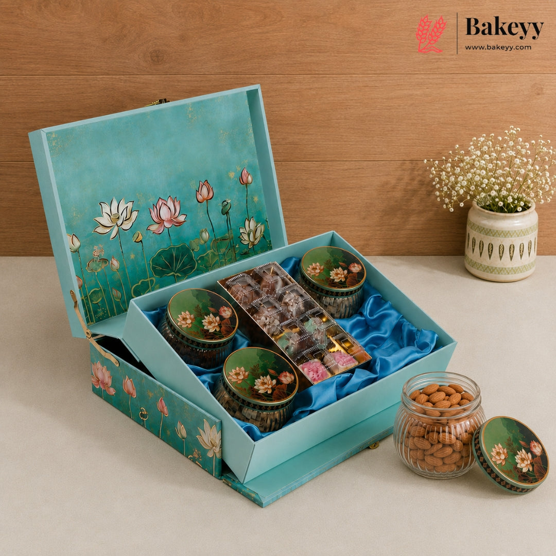 A teal and pink lotus-themed gift box from Bakeyy, filled with assorted chocolates and sweets, displayed on a beige surface against a wooden background with a small potted plant.