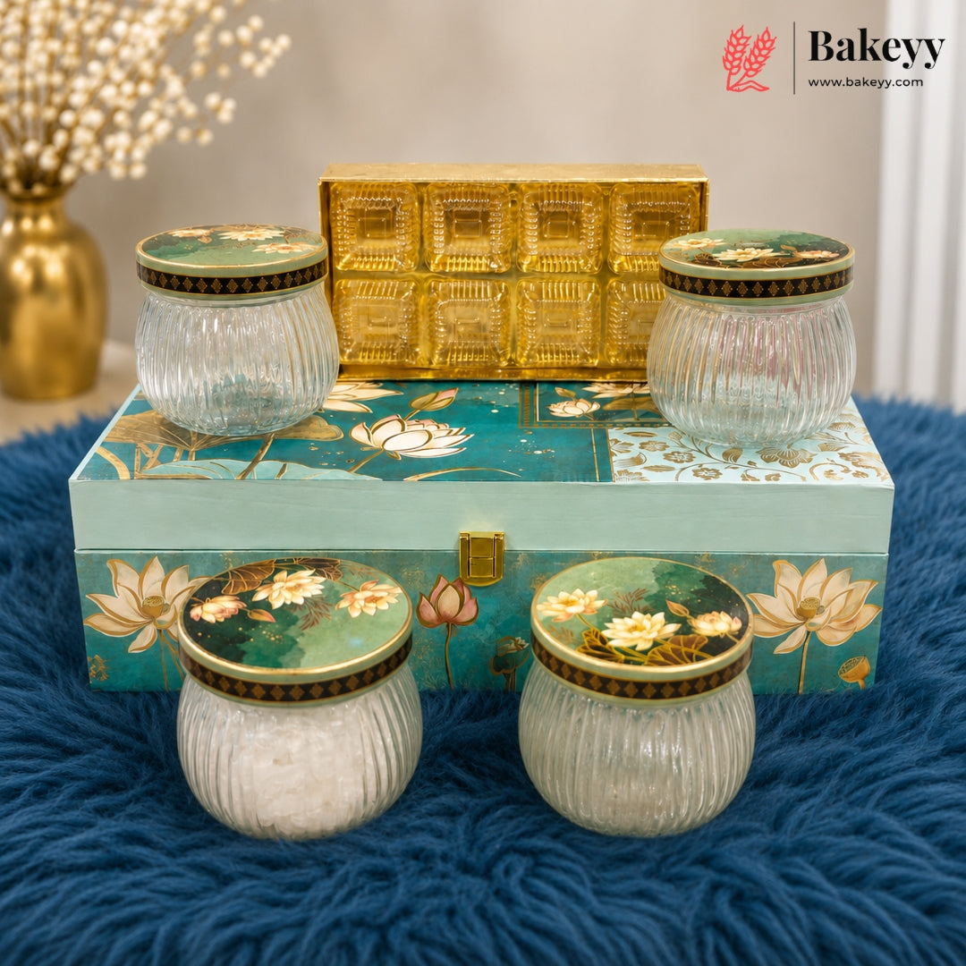 A set of five decorative glass jars with ribbed clear bodies and floral-patterned lids, arranged on and around a painted teal box with a gold latch, plus a small gold decorative box behind them. The scene is styled on a blue textured surface with a blurred vase in the background.