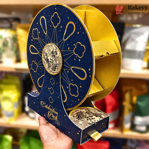 A hand holds a decorative round gift box with a navy blue and gold design, featuring cloud motifs, a central moon emblem, and the words 'Celebration Pack' on a blurred store shelf background.
