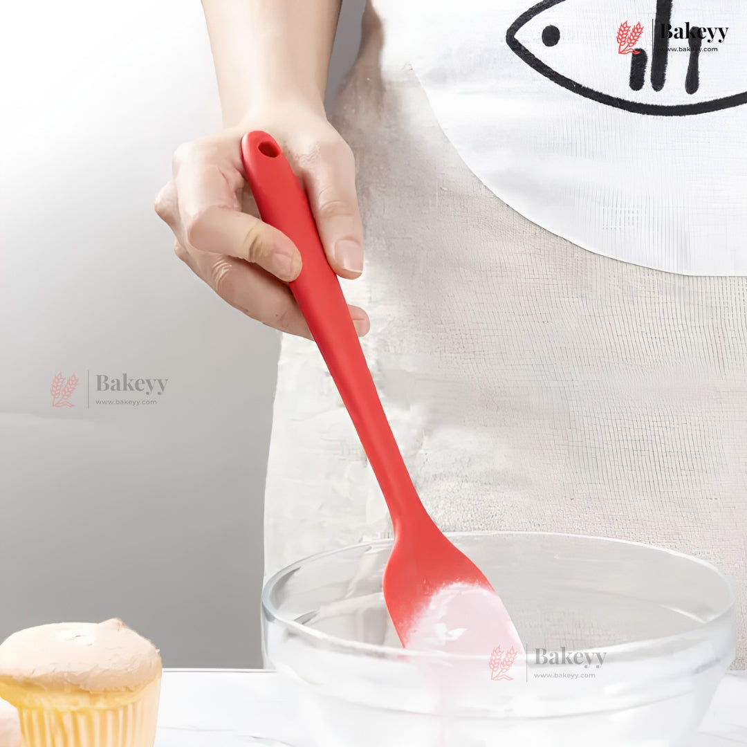 Red silicone spoon held by a person with a white apron, near a glass bowl and cupcake.