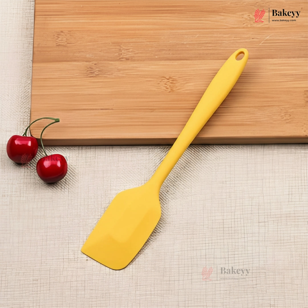 Yellow spatula on a wooden cutting board with two red cherries, on a beige surface with 'Bakeyy' branding.