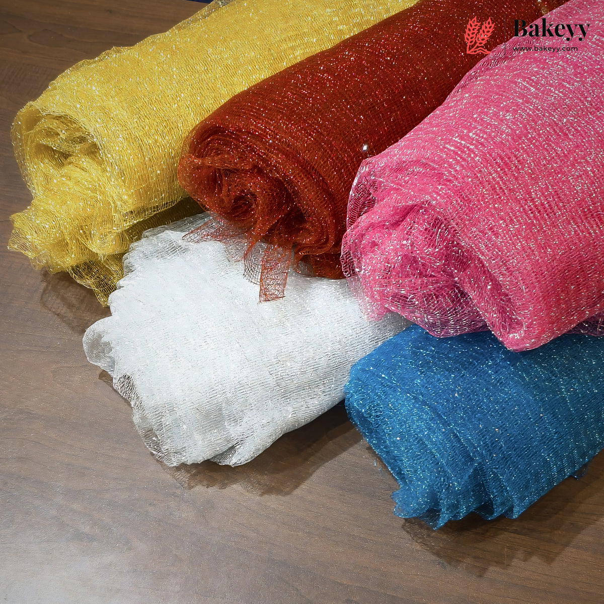 Five rolls of sparkly tulle mesh fabric in yellow, rust red, white, pink, and blue, arranged diagonally on a dark wood surface. A 'Bakeyy' brand logo is visible in the top right corner.