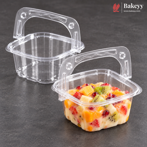 Two clear plastic square food containers with integrated handles, one empty and one filled with a fruit yogurt dessert, on a dark gray surface. The Bakeyy logo is visible in the top right corner.