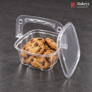 A clear plastic bakery cookie container with a handle and lid, shown open and filled with chocolate chip cookies on a dark gray surface. The Bakeyy logo is visible in the top right corner.