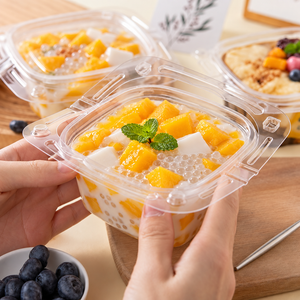 A close-up of a hand holding a clear plastic cup of dessert with mango chunks, white dessert pieces, tapio pearls, and a mint leaf on top. Another similar dessert container is blurred in the background on a wooden surface.