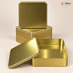 Three gold-colored metal tin boxes arranged on a light beige background, with one open box in the foreground and two stacked closed boxes behind it. A small Bakeyy logo appears in the top right corner.