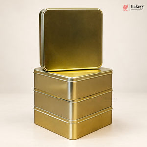 Three stacked square metal tin boxes in a metallic gold finish, with the top tin slightly offset to show the lid, photographed on a clean light beige studio background. A Bakeyy watermark/logo appears in the top right corner.