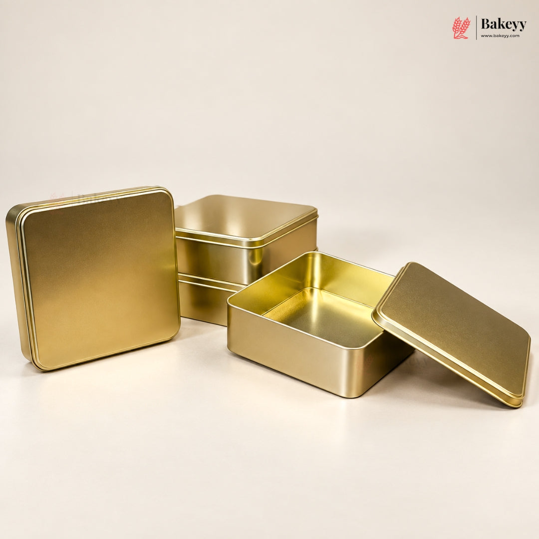 Three square gold metal tin boxes with lids on a clean off-white background, with one box open to show the interior. A small Bakeyy logo appears in the top right corner.