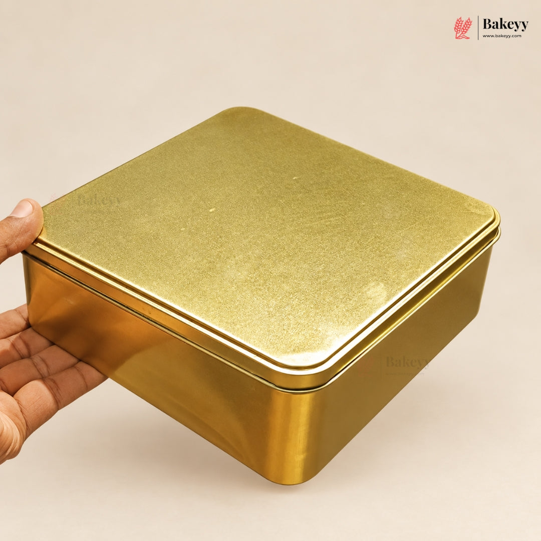 A square metallic gold tin container with a hinged lid, held by a hand on the left side against a plain light beige background. A Bakeyy logo watermark is visible in the top right corner.