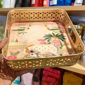 A square decorative tray with a gold-toned perforated metal border and a colorful printed inner design featuring a white spotted cow, pink lotus flowers, green leaves, and domed architecture. A hand is holding the tray in a retail setting.