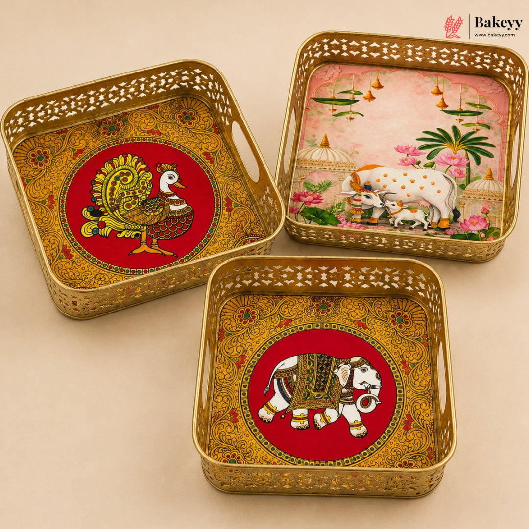 Three decorative square metal trays with perforated edges, arranged on a beige background. Each tray features a red circular center with traditional Indian-style artwork: one shows a peacock, one shows a cow and calf, and one shows a white horse.