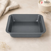 A dark gray rectangular non-stick baking pan with raised edges, shown on a light beige countertop with a folded cloth, a bowl of flour, and a whisk nearby. The Bakey logo appears in the top right corner.