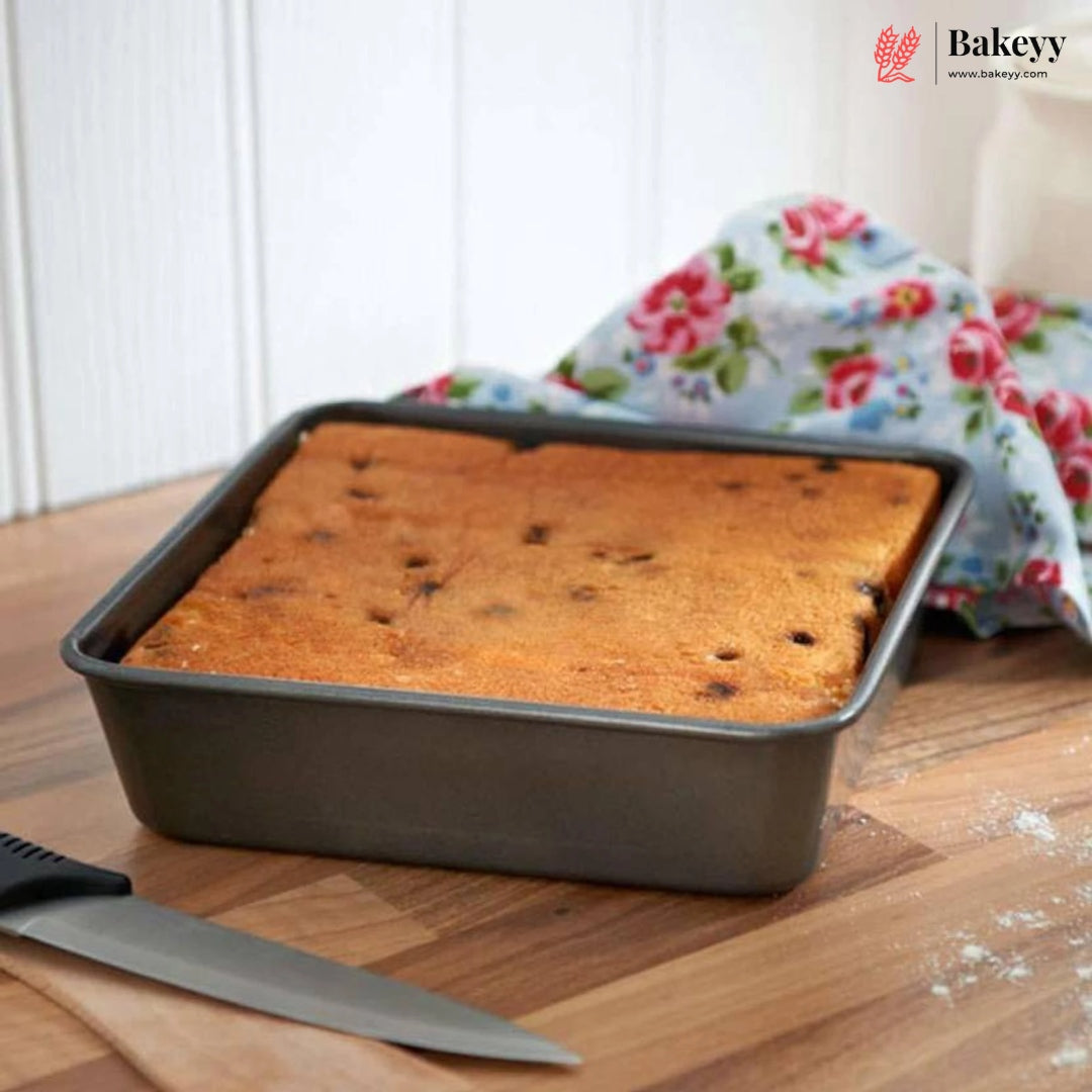 A rectangular dark gray baking pan containing a golden-brown rectangular cake, set on a wooden surface with a floral cloth and a knife nearby. The Bakey brand logo is visible in the top right corner.