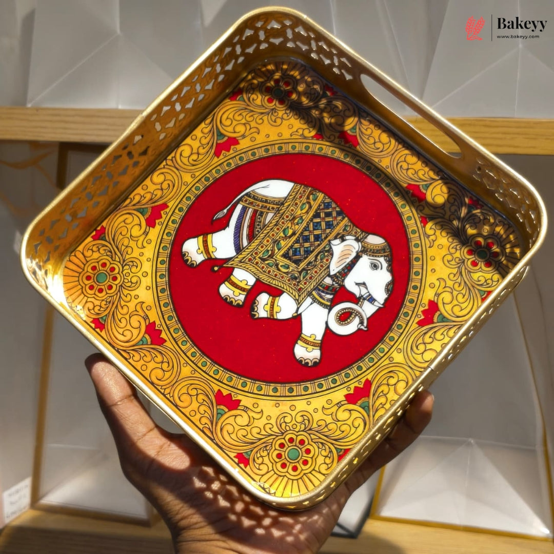 A square decorative tray with cutout handles, held in a hand. The tray features a vibrant red and gold background with an intricate white elephant illustration in the center.