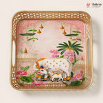 A square decorative serving tray with a gold-toned perforated border and integrated side handles, featuring a hand-painted illustration of a white cow and calf among pink lotus flowers, green leaves, hanging bells, domed pavements, and a soft pink sky. The Bakeyy logo appears in the top right corner.
