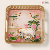 A square decorative serving tray with a gold-toned perforated border and integrated side handles, featuring a hand-painted illustration of a white cow and calf among pink lotus flowers, green leaves, hanging bells, domed pavements, and a soft pink sky. The Bakeyy logo appears in the top right corner.