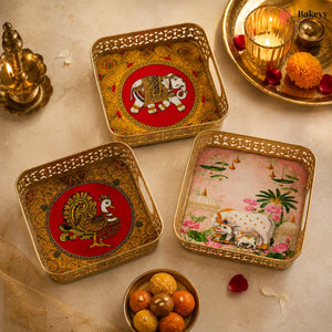 Three decorative square metal trays with ornate cutout edges, arranged on a beige surface with festive props. Each tray has a gold-toned border and a central painted motif: one red circle with an elephant, one red circle with a peacock, and one pink scene with cows and flowers.