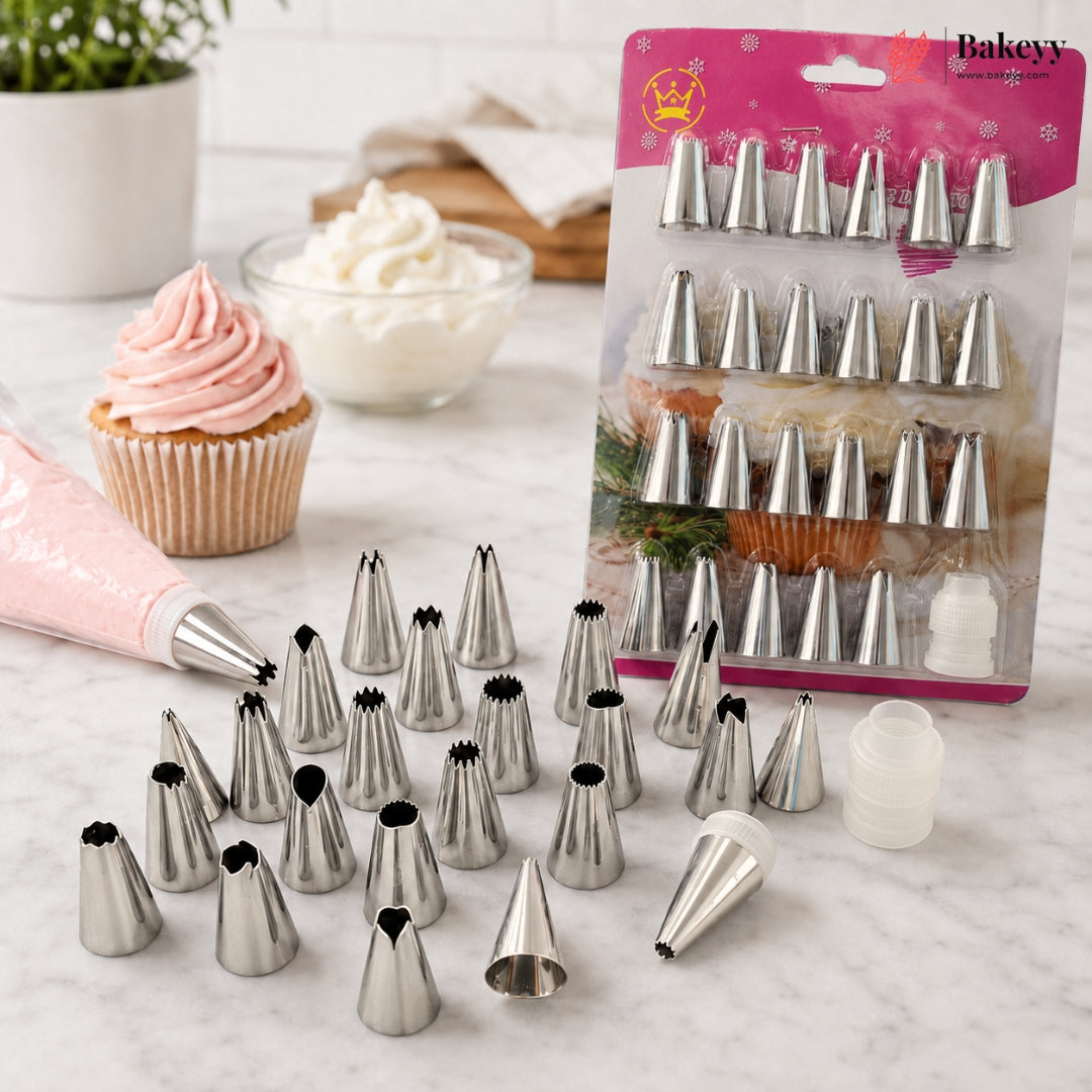 A set of metal piping tips displayed on a marble surface, with a pink piping bag and a packaged set of baking tips in the background. A cupcake with pink frosting and bowls of frosting are also visible.