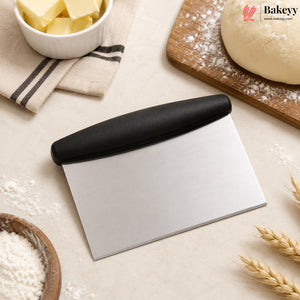 A stainless steel pastry scraper with a black handle, styled on a beige surface with baking ingredients including flour, butter, dough, and wheat stalks. The Bakey brand logo is visible in the top right corner.