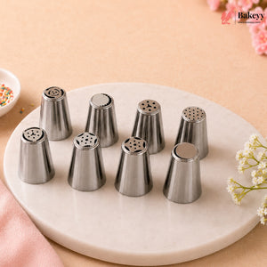 Eight stainless steel piping nozzles arranged on a round white marble tray against a warm beige background with soft pink floral accents. A Bakeyy logo appears in the top right corner.
