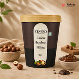 A 1kg tub of TENERO Choco Hazelnut Filling, featuring a dark brown label with a cream-colored center label and a gold lid. The tub is styled on a beige stone surface with hazelnuts, chocolate pieces, and a small bowl of the chocolate filling, under soft, warm lighting.