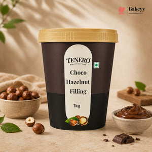 A 1kg tub of TENERO Choco Hazelnut Filling, featuring a dark brown label with a cream-colored center label and a gold lid. The tub is styled on a beige stone surface with hazelnuts, chocolate pieces, and a small bowl of the chocolate filling, under soft, warm lighting.