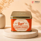 A square metal tin of Tiger Kesari Colour food coloring, displayed on a round beige pedestal. The tin has a metallic gold lid and a bright orange label with a white central badge.