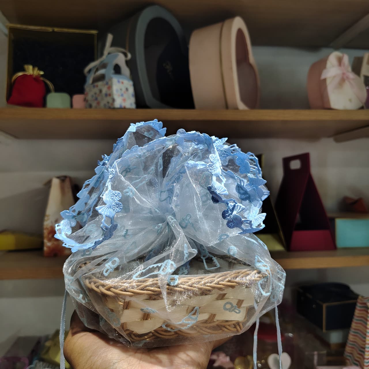 Hand holding a small woven basket with decorative blue lace over a blurred background of shelves with various items.