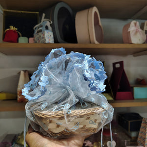 Hand holding a small woven basket with decorative blue lace over a blurred background of shelves with various items.