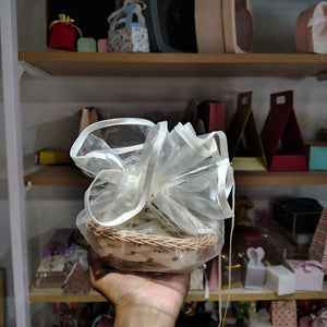 Hand holding a gift basket with a large white bow in front of a shelf with various items.