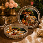 Two ornate decorative plates with intricate silver borders, featuring a painted scene of decorated cows under a tree. One plate is open, styled with a small brass idol, flowers, and a lit candle, while the other stands closed on a wooden surface with a brass lamp and flowers in the background.
