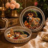Two ornate decorative plates with intricate silver borders, featuring a painted scene of decorated cows under a tree. One plate is open, styled with a small brass idol, flowers, and a lit candle, while the other stands closed on a wooden surface with a brass lamp and flowers in the background.